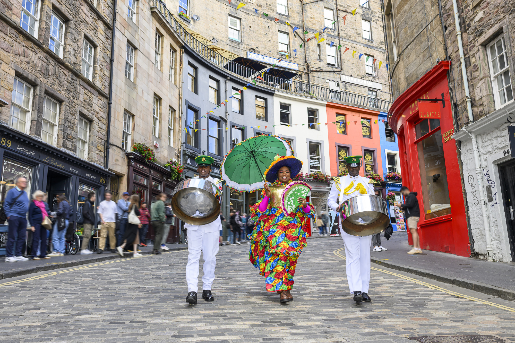 Festival performers in traditional dress walking down a street carrying steel drums and a parasol