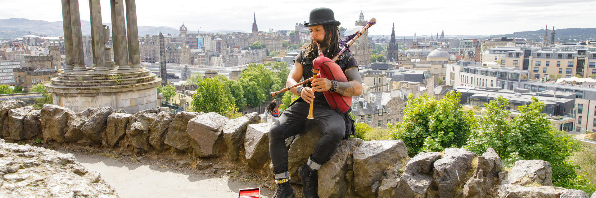 Dudelsackspieler sitzt auf einer Steinmauer mit Stadtblick und klassischem historischen Denkmal links.