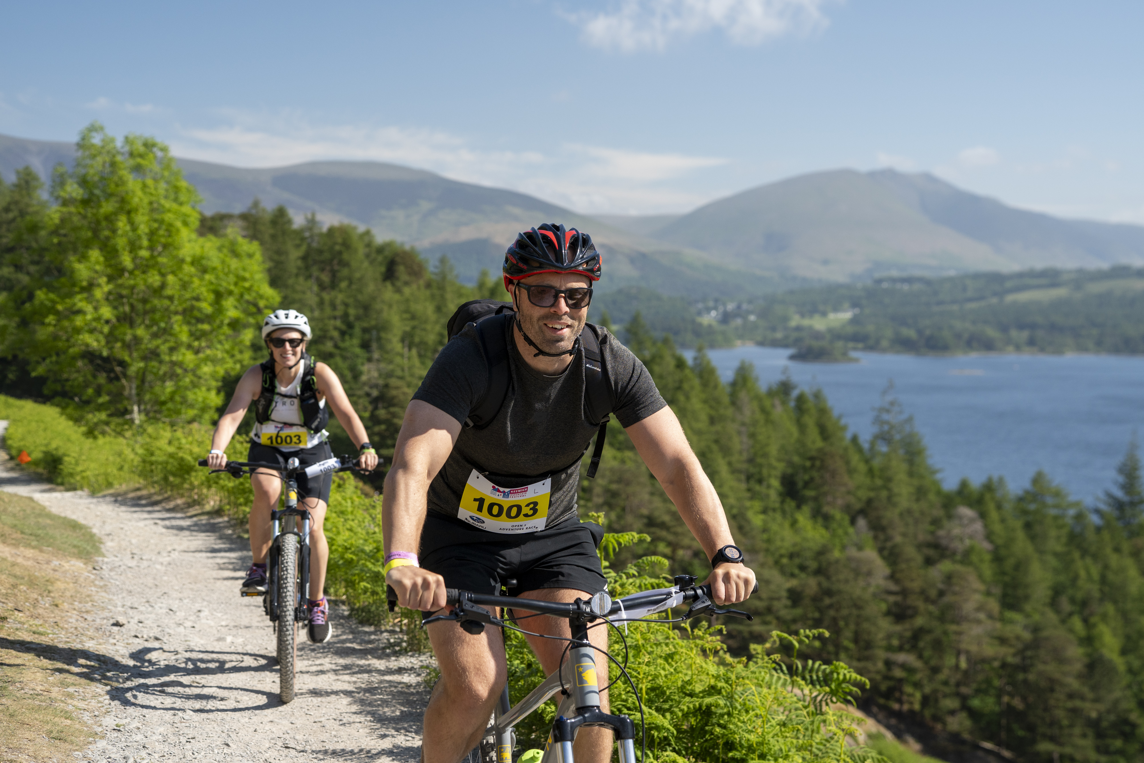 Two cyclists ride on a scenic trail beside a lake, surrounded by green hills and distant mountains under a blue sky.