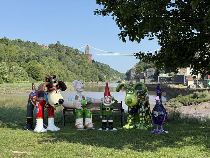 Sculptures of animated characters on grass near a river, with a suspension bridge and green hills in the background on a sunny day.