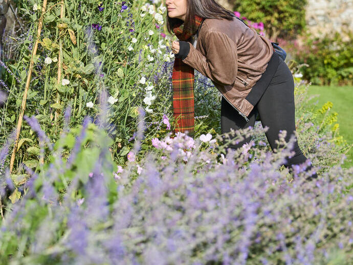 Mujer huele flores en un jardín junto a un muro de piedra.