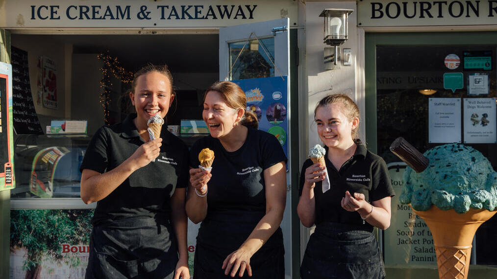 Three smiling woman standing outside ice cream parlour.