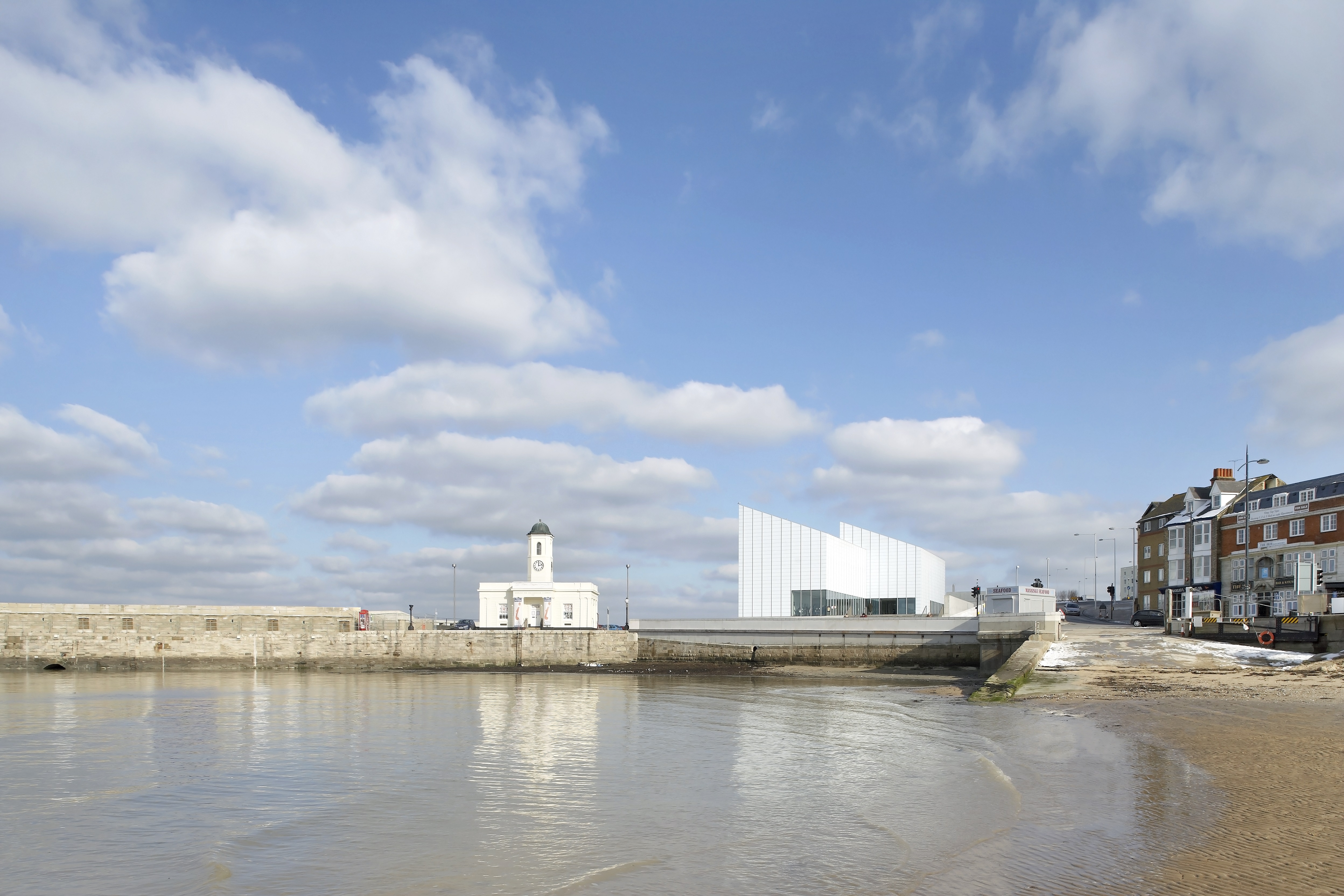 Modern building overlooking a harbour and beach, a row of townhouses in the background