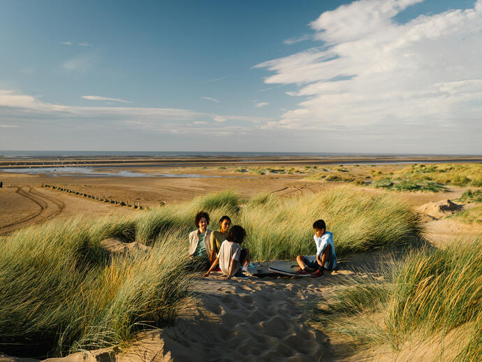 Four people sit on sand dunes among tall grass, with a wide sandy beach and tidal flats extending to the horizon under a blue sky.