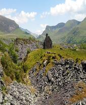 Ruined stone building on grassy hill, surrounded by slate and scattered rocks, with mountains and blue sky in the background.