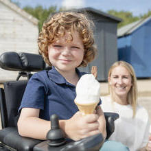 Young electric wheelchair user holding an ice cream cone with mum in background at the seaside