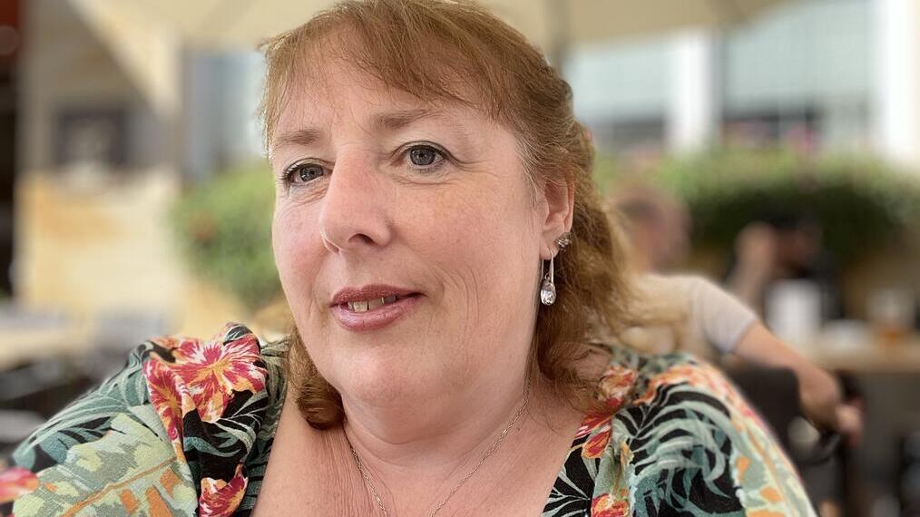 Woman with floral shirt seated outdoors under an umbrella, with blurred background of plants and people at a café or restaurant.