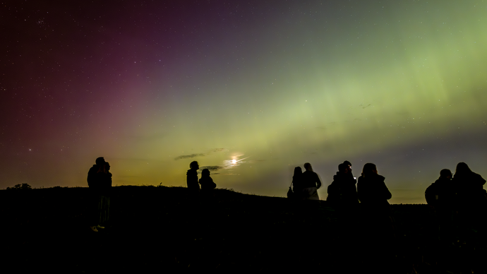 Silhouetted group of people watching the northern lights in a dark landscape under a colorful night sky.