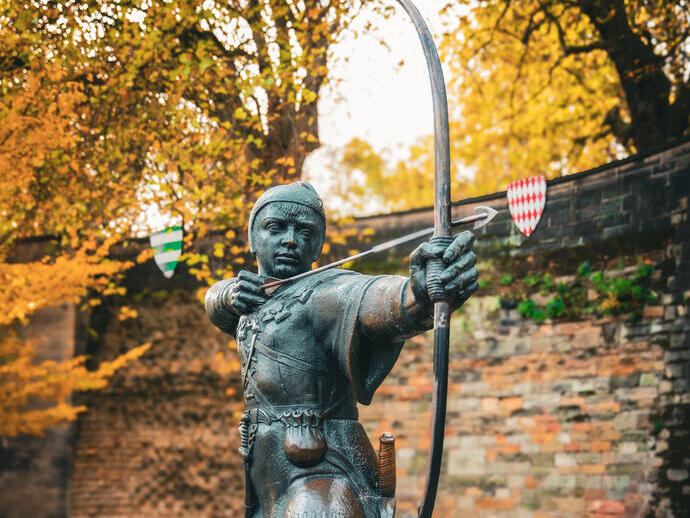 Statue of an archer drawing a bow, set outdoors with autumn trees and a stone wall in the background.