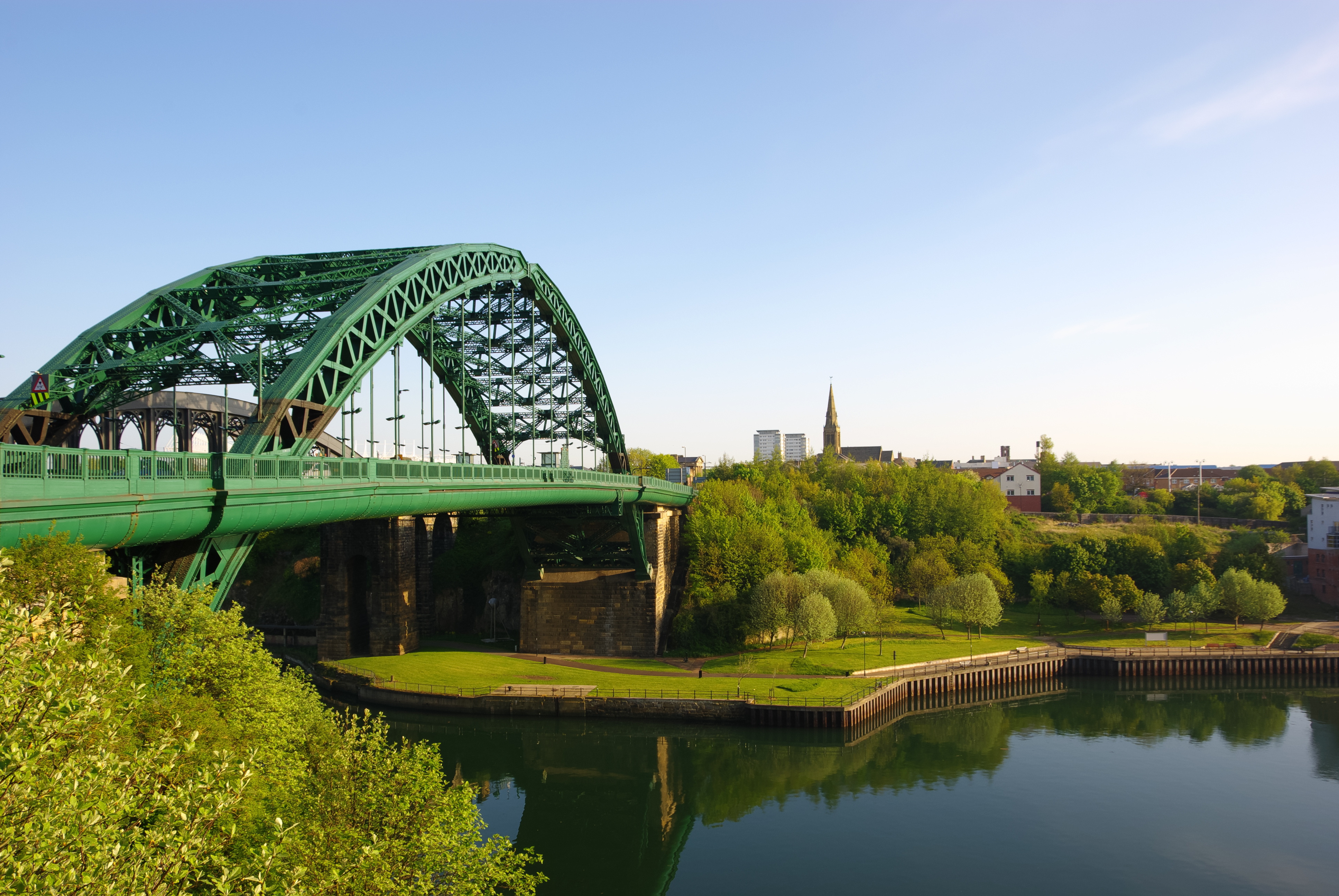 View of a green iron bridge crossing over a river, surrounded by trees and buildings