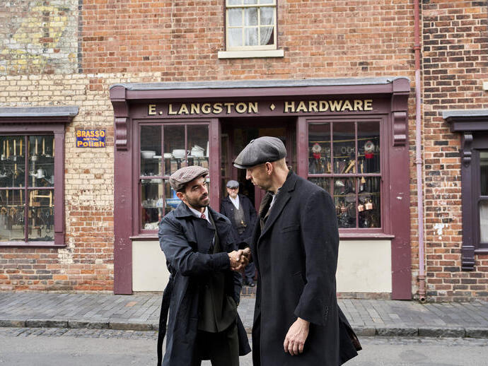 Two men in vintage attire shake hands outside a brick hardware store named E. Langston. Historical street scene with older shopfronts.