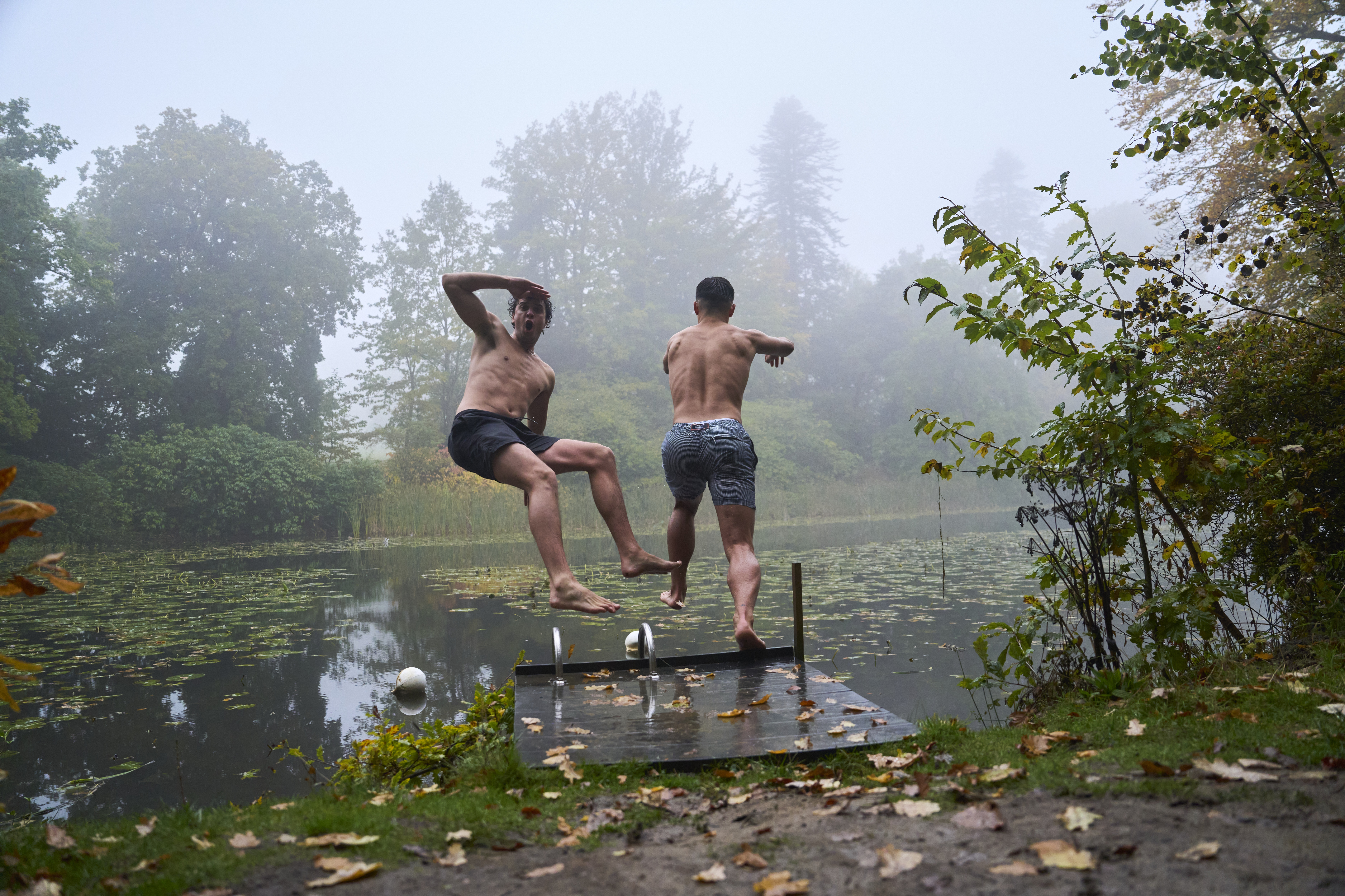 Zwei Männer springen von der Badeplattform in einen See in herbstlicher Umgebung
