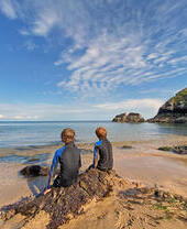 Back view of two young boys in wetsuits sitting on a rock looking out to sea with blue sky