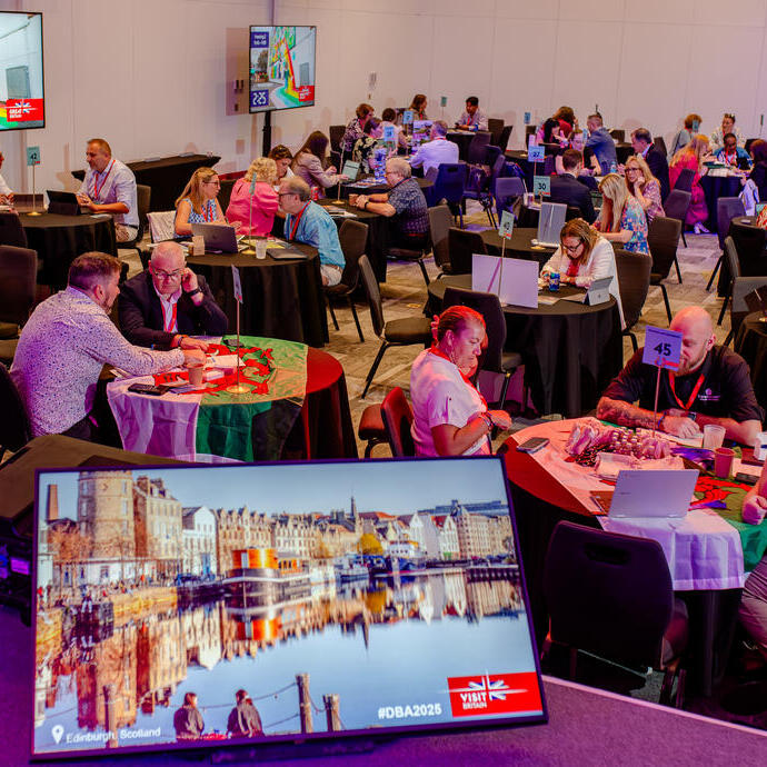 A conference room with round tables, people networking, laptops open, and a screen showing an image of Edinburgh, Scotland. Professional event atmosphere.