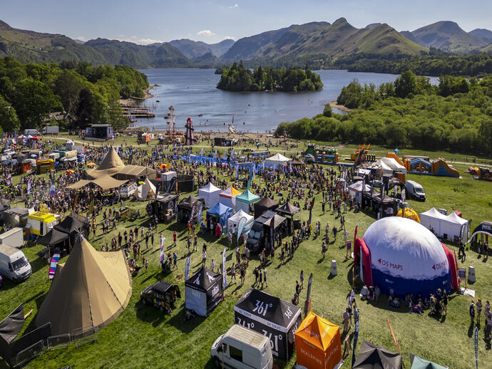 Outdoor festival with tents, food trucks, and crowds by a lake, surrounded by mountains and lush greenery on a sunny day.