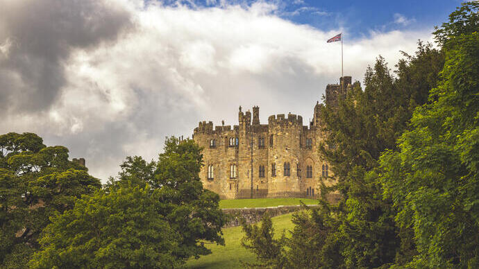 Historic stone castle on a hill, partially surrounded by lush green trees, under a cloudy sky with a flag atop the building.