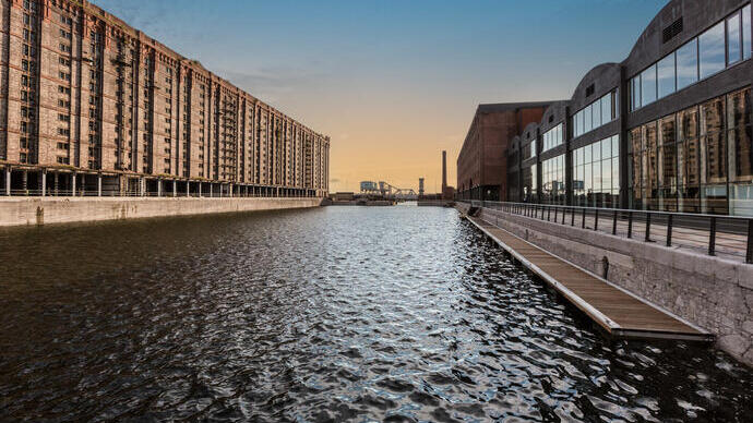A view of a dock looking across the water. To the left stands a traditional brick built dockside building, to the right is a modern building made from glass and wood.