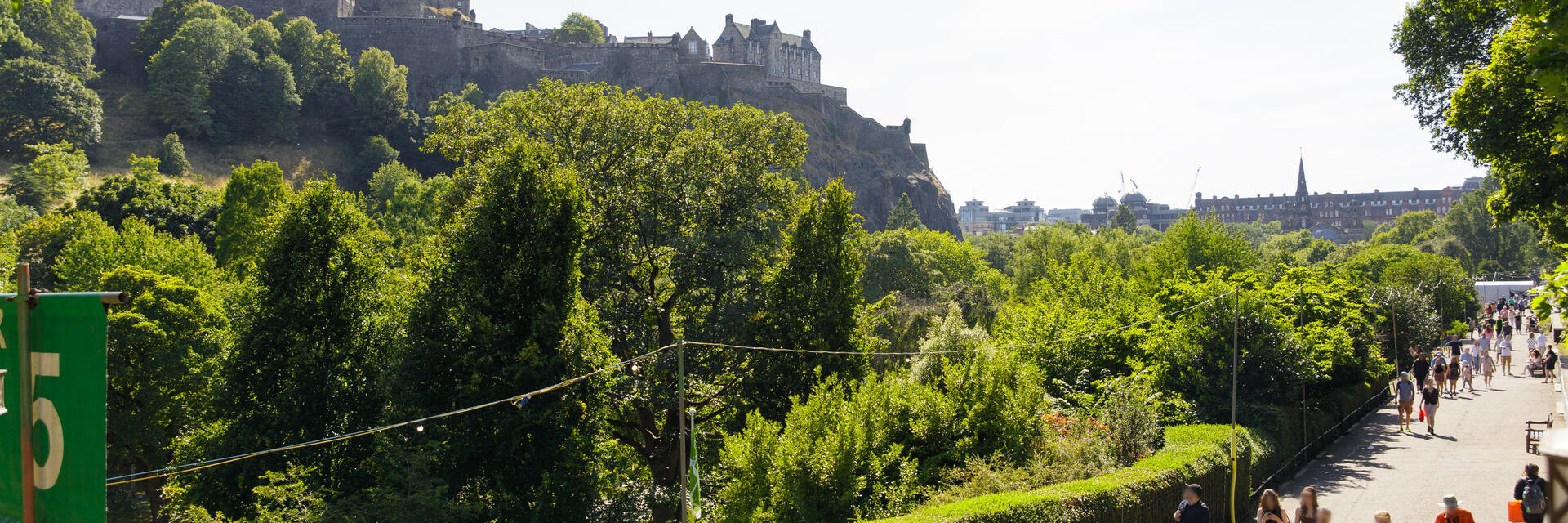 Blick auf das Edinburgh Castle, umgeben von Bäumen, mit Menschen auf einem sonnigen Weg im Vordergrund.