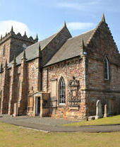 Historic stone church with pointed gables and clock tower, surrounded by gravestones in a rural landscape under a blue sky.