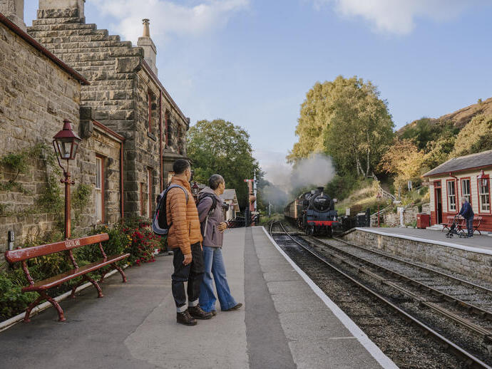 Stazione ferroviaria rurale in pietra con passeggeri in attesa sul binario e treno a vapore in arrivo. Circondata da alberi ed edifici tradizionali.