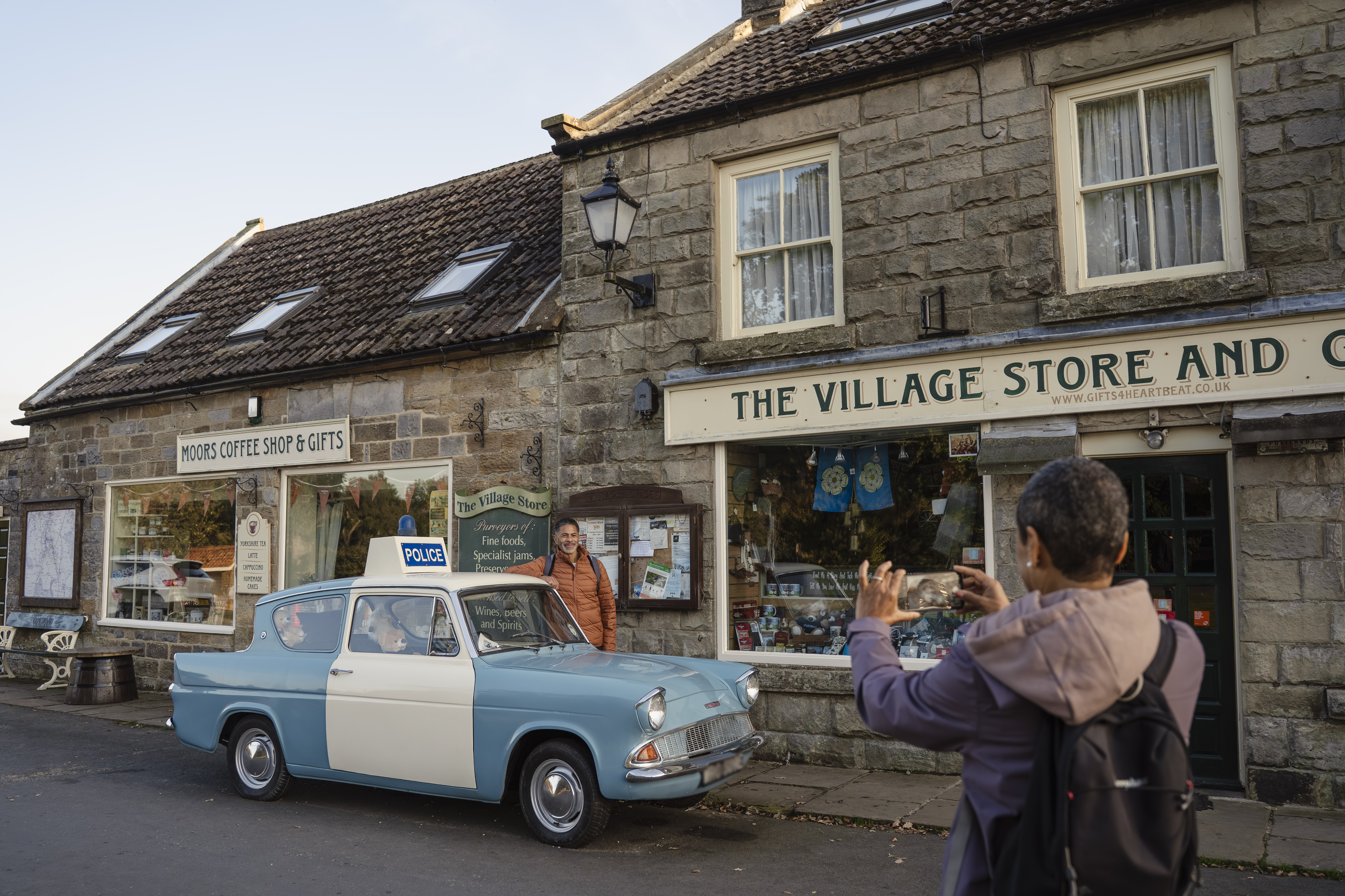 Street view of two people and a vintage police car outside a stone village store and coffee shop, with gift shop signs.