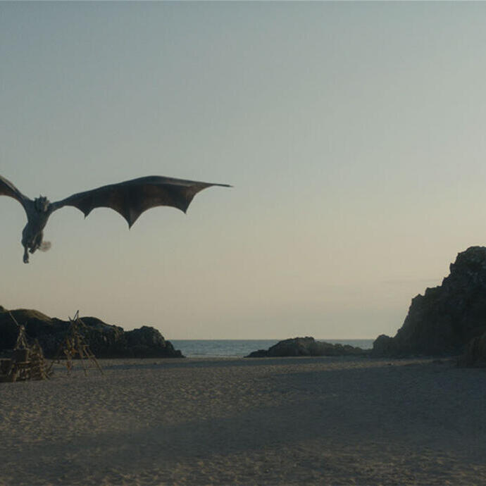 A large dragon flies over a sandy beach with rocky outcrops and the sea in the background, under a clear sky.