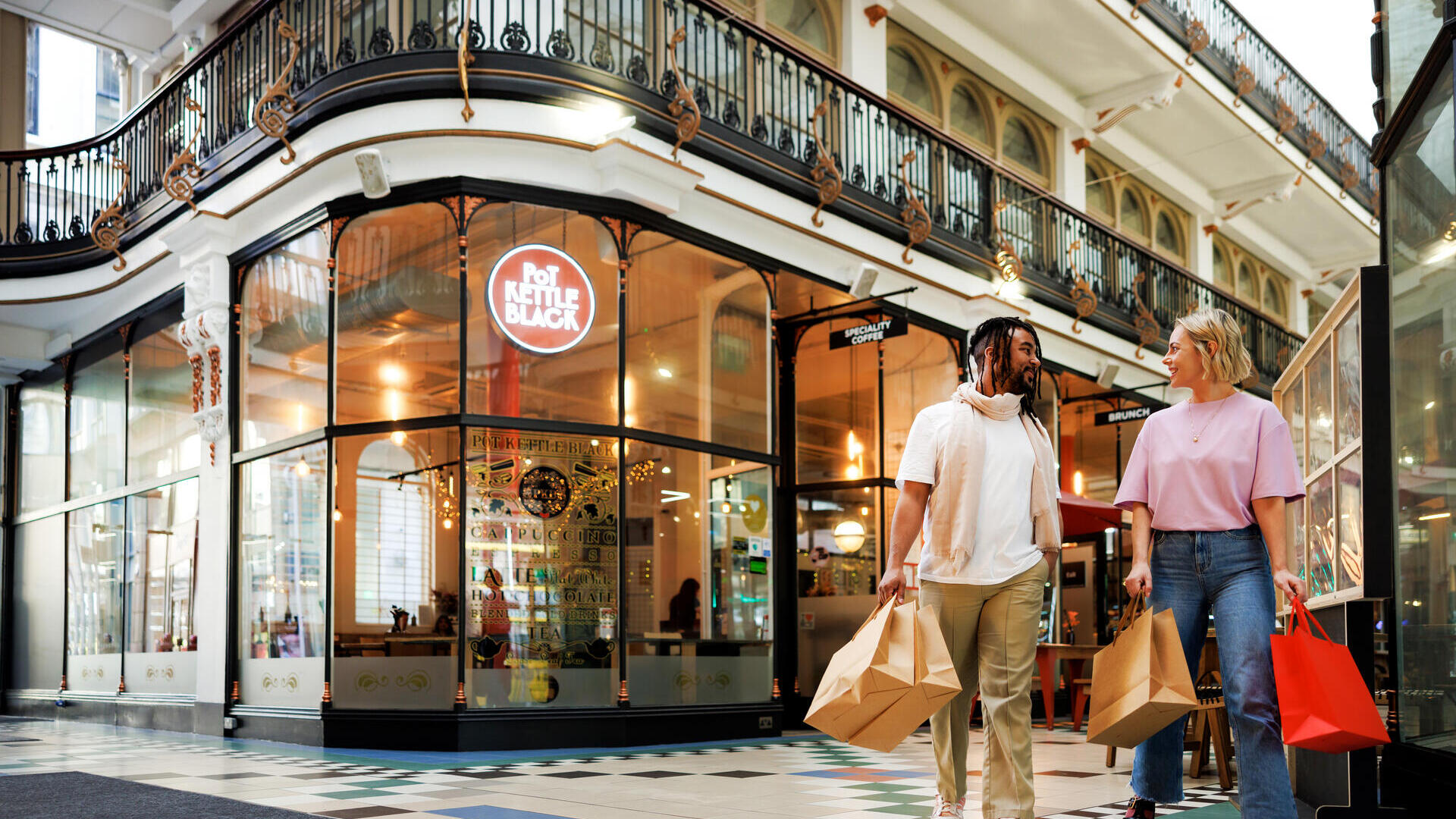 Two people carrying shopping bags walk through a stylish indoor arcade with ornate railings and a café in the background.