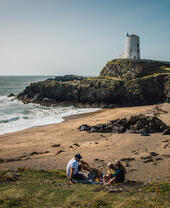 Une jeune famille pique-niquant sur une plage avec un phare en arrière-plan.