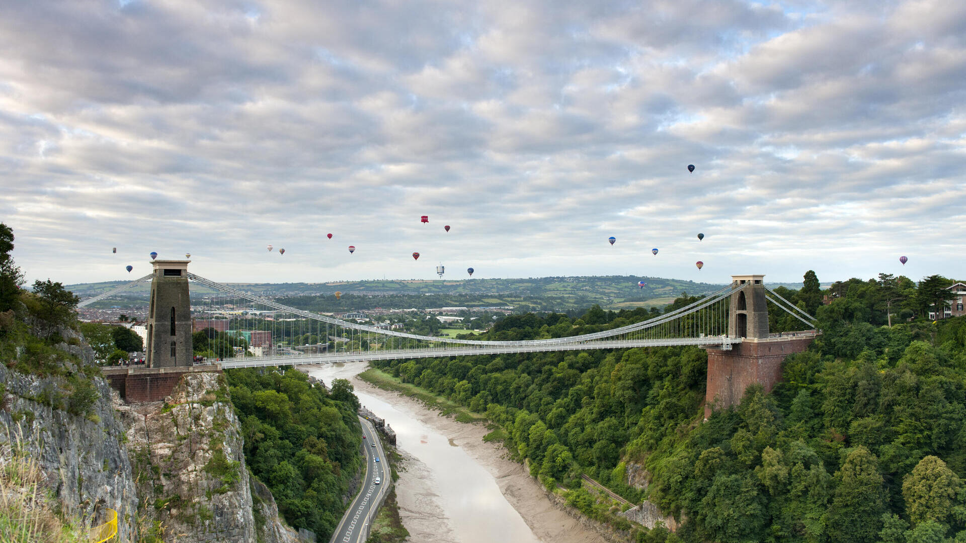 Hot air balloons floating above a river and a suspension bridge.