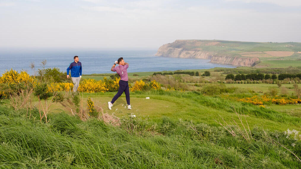 Deux hommes jouent au golf sur un parcours avec la côte et le promontoire en arrière-plan.