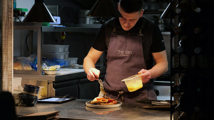 Chef dans une cuisine professionnelle concentré sur le dressage d'un plat, portant un tablier, entouré d'ustensiles de cuisine et d'ingrédients préparés.