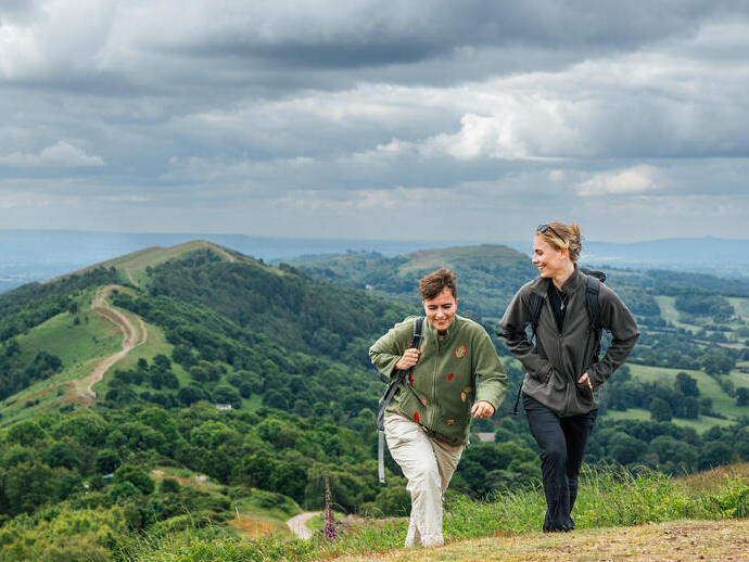 Two women hike carrying rucksacks along green hills on a cloudy day