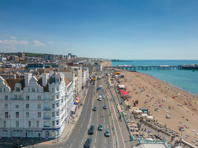A view of a busy seaside city with a sandy beach, pier, seafront road, historic buildings, and people enjoying a sunny day.