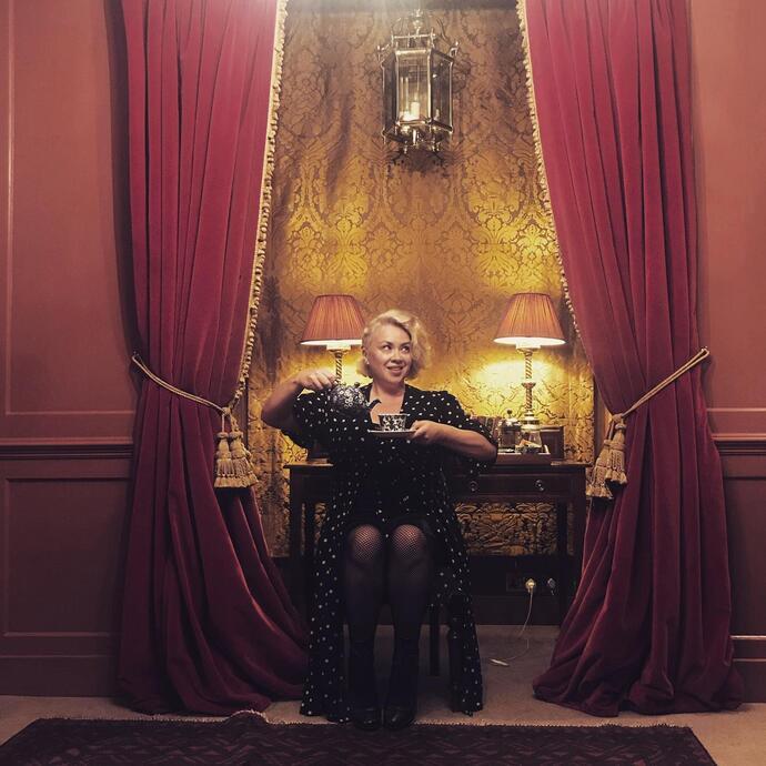 A woman sits on a chair pouring a cup of tea in front of red velvet curtains at the Hazlitt's Boutique Hotel in Soho London