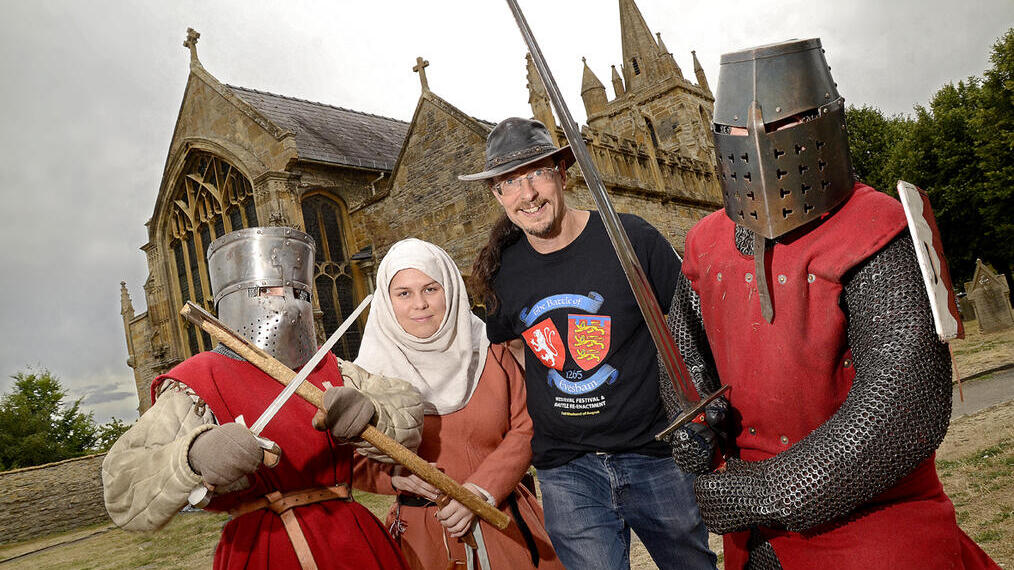 Four people in medieval costumes, including two in chainmail and helmets, pose with swords outside a historic church.