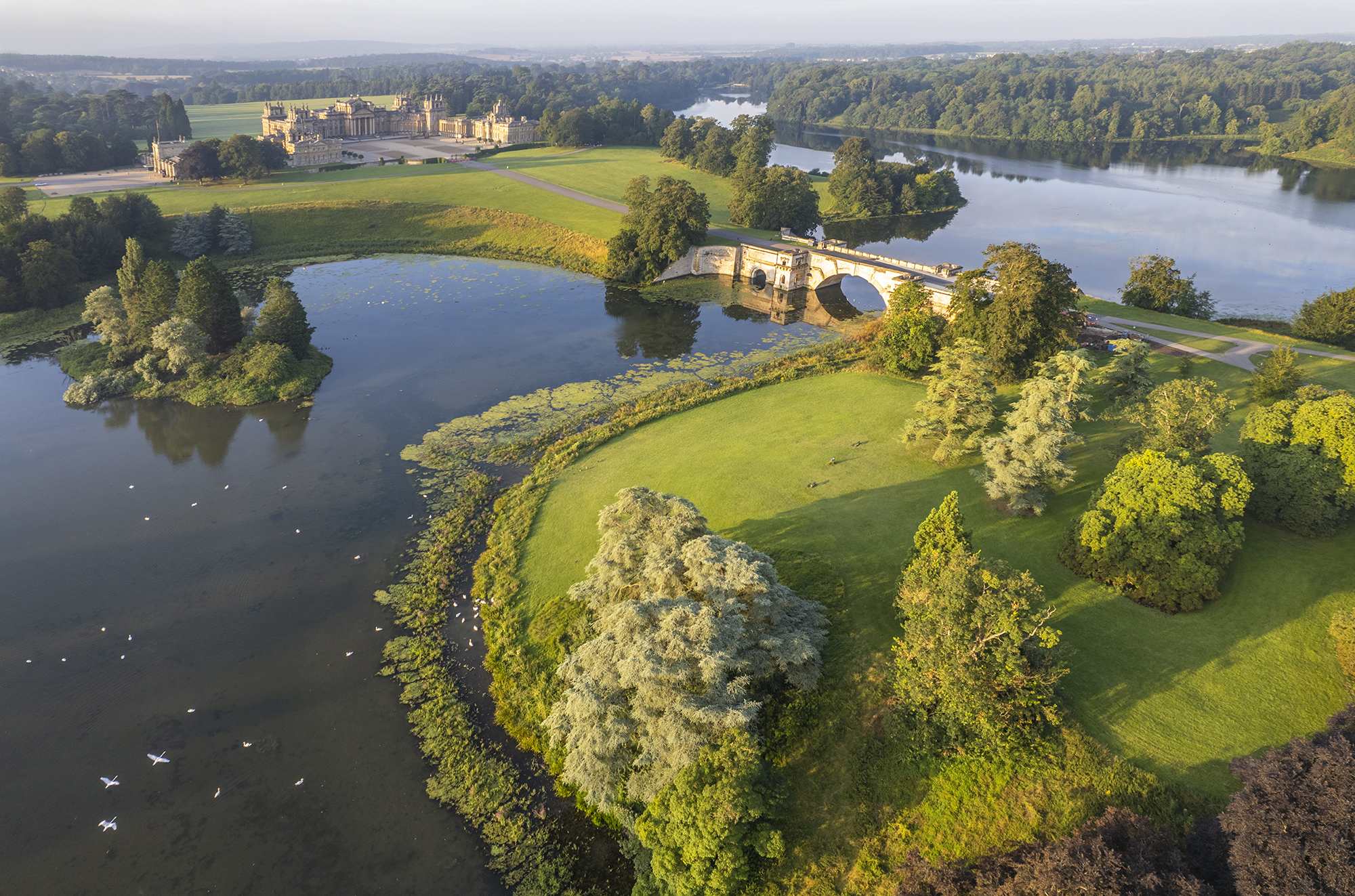 Vista aérea de un palacio del siglo XVIII, rodeado de jardines, árboles y un lago atravesado por un puente.