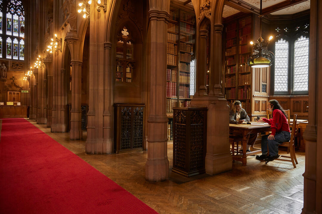 Two men chatting, seated inside a large library reading room.