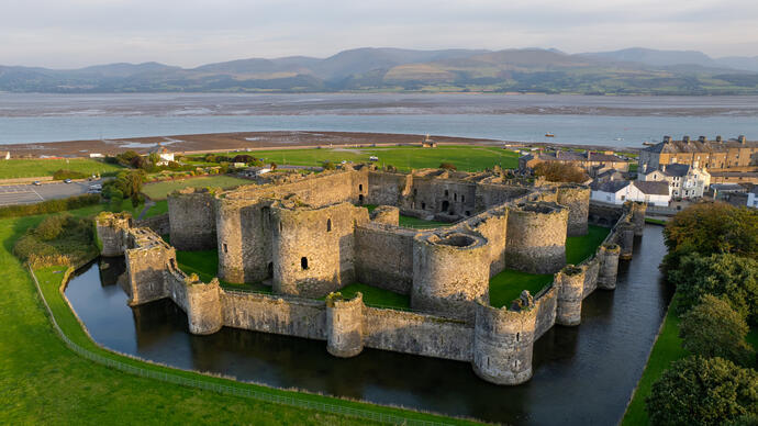 Aerial view of a historic stone castle with round towers, surrounded by a moat, near a coast, with mountains in the background.