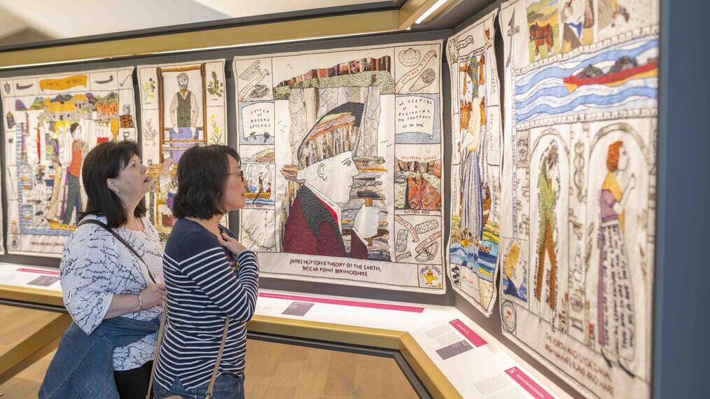 Two people viewing a display of embroidered panels in a museum setting, with illustrated historical and geological themes.