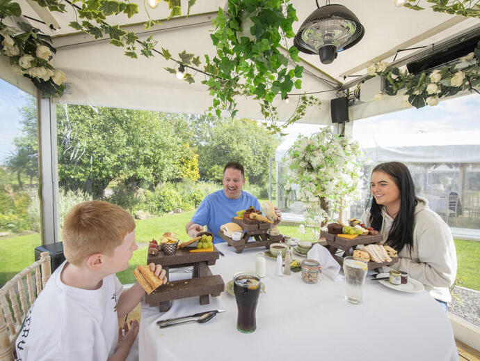 Famiglia che mangia insieme in un gazebo esterno decorato con piante e fiori, tavolo imbandito e giardino sullo sfondo.