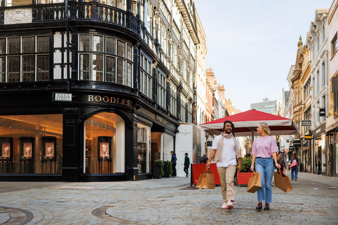 Two people carrying shopping bags walk on a cobbled street with historic buildings and shopfronts, including Boodles, in view.