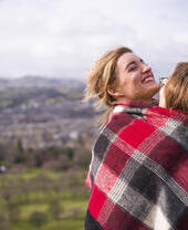 Two women wrapped in red checkered blanket standing on a hill overlooking a city