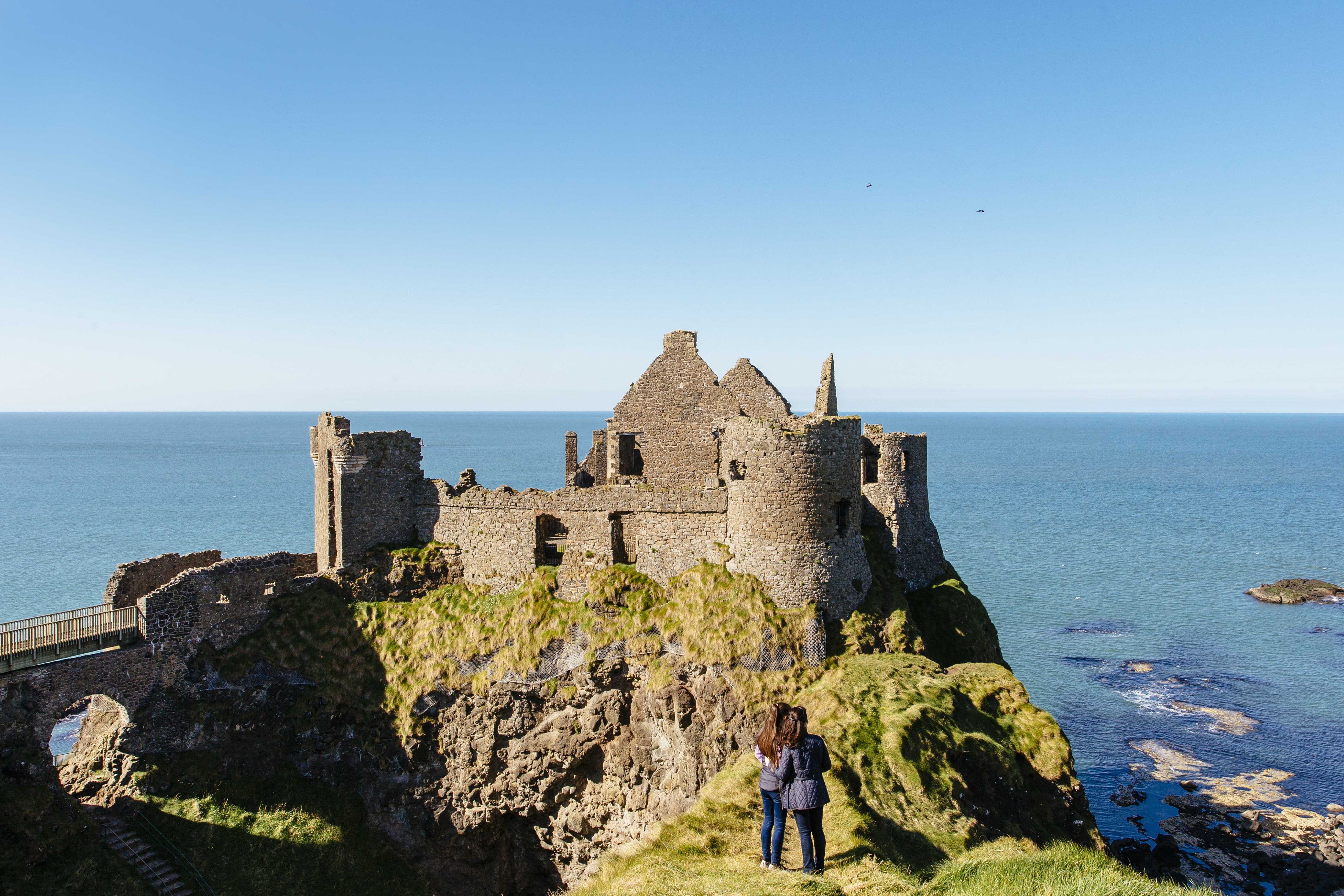People looking at an historic stony castle from a rocky outcrop cliff over the ocean.