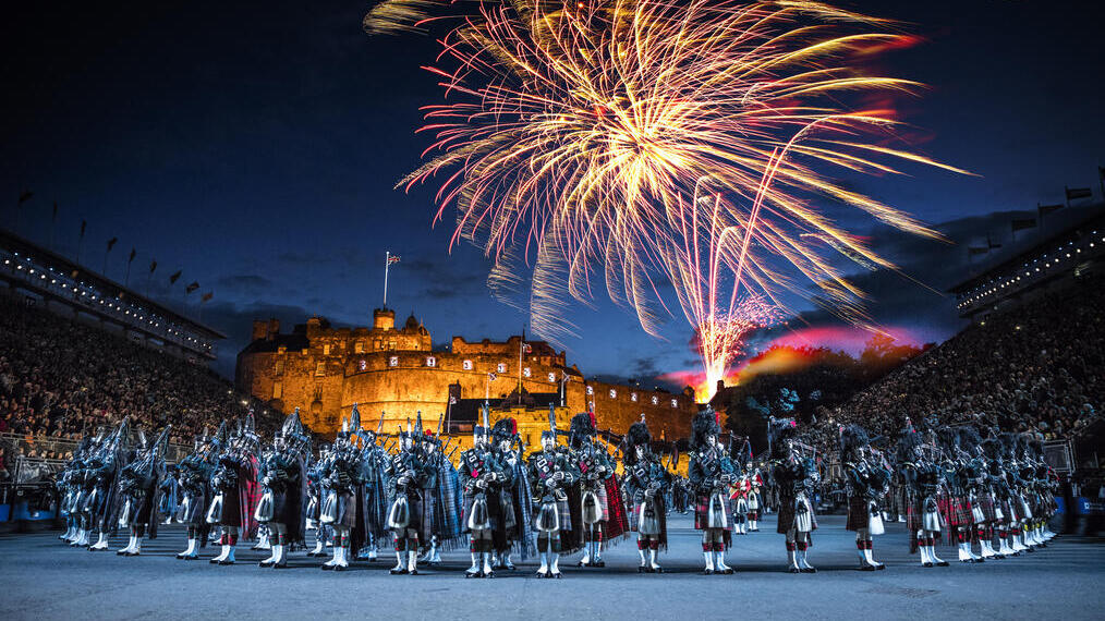 The arena at the Edinburgh Military Tattoo during a performance of the military event, parade ground and packed spectator seating. A light show projecting onto the castle walls. Marching band with a leading conductor, and massed pipers playing the bagpipes. Fireworks exploding in the night sky.