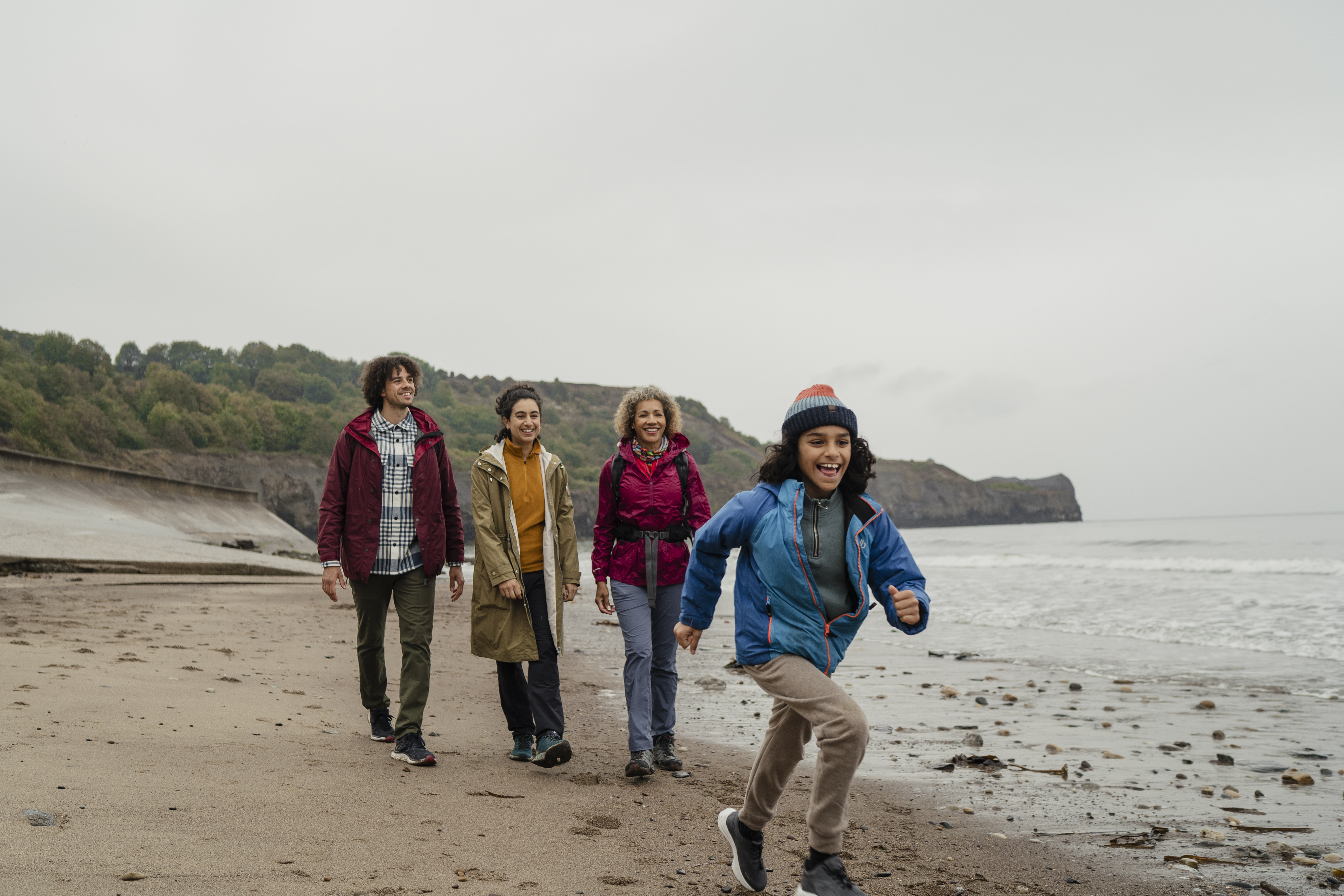 عائلة تسير على شاطئ رملي بينما يركض طفل أمامهم، مع منحدرات وخلفية تغطيها الأشجار.