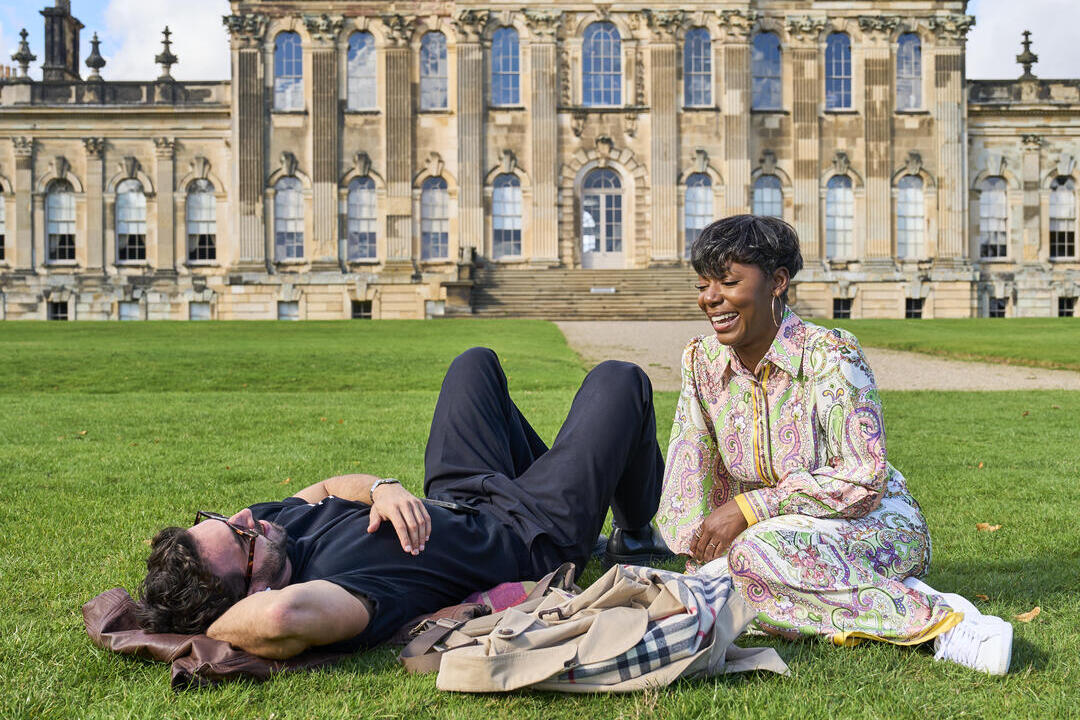Un homme et une femme détendus et souriants sur l’herbe devant un grand bâtiment historique avec colonnes et dôme.