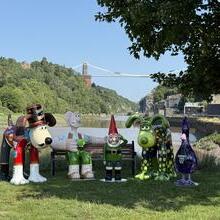Sculptures of animated characters on grass near a river, with a suspension bridge and green hills in the background on a sunny day.
