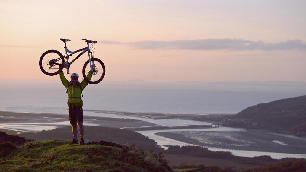 Man holding his mountain bike above his head in celebration at the top of a hill with the coast below him