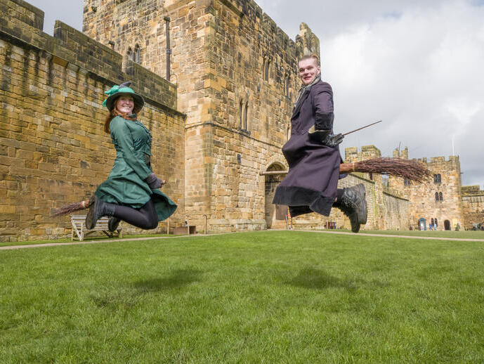 Two wizarding professors demonstrating flying on a broom outside a large castle.