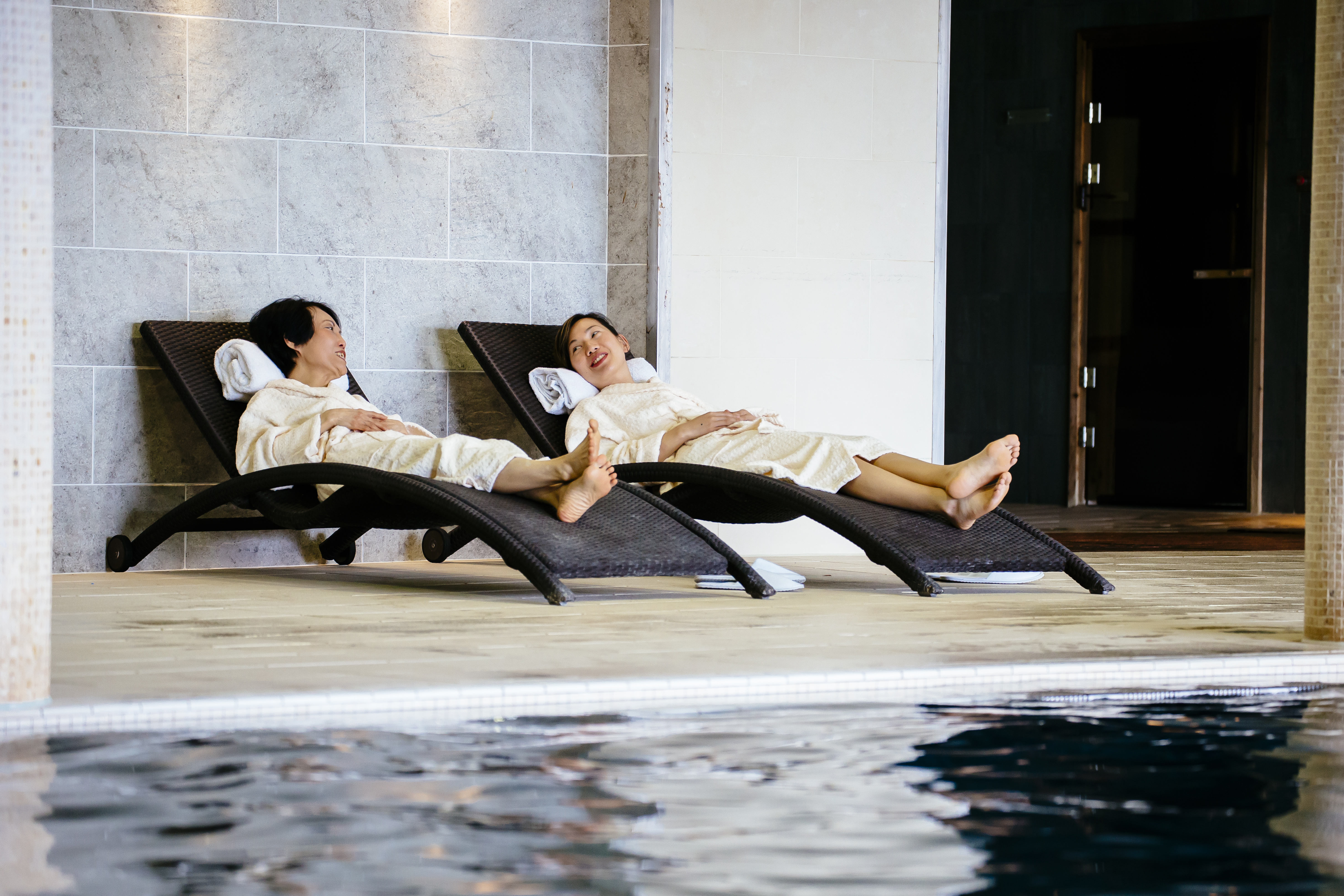 Two women lying on loungers beside an indoor swimming pool